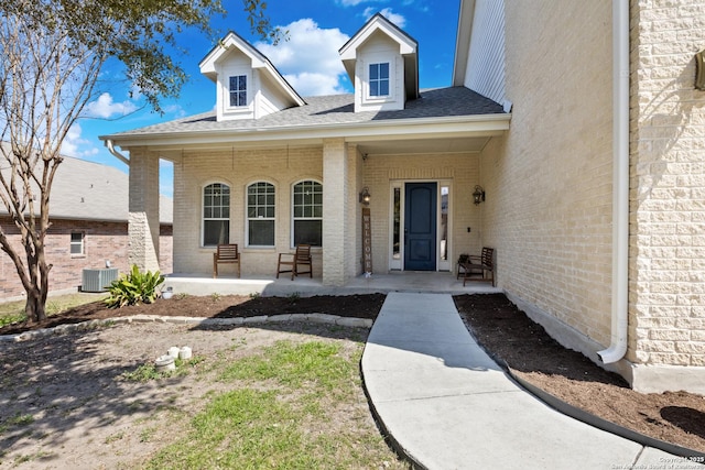 entrance to property with brick siding, a porch, central AC unit, and a shingled roof