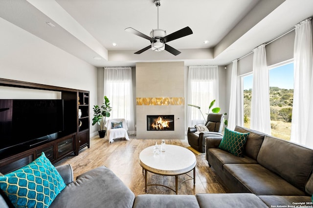 living room featuring a ceiling fan, a tile fireplace, a raised ceiling, and recessed lighting