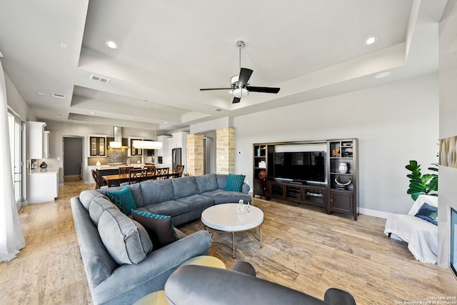 living room with baseboards, visible vents, a raised ceiling, ceiling fan, and light wood-style floors