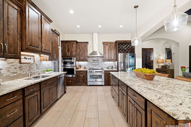 kitchen featuring light stone countertops, stainless steel appliances, dark brown cabinets, wall chimney range hood, and a sink