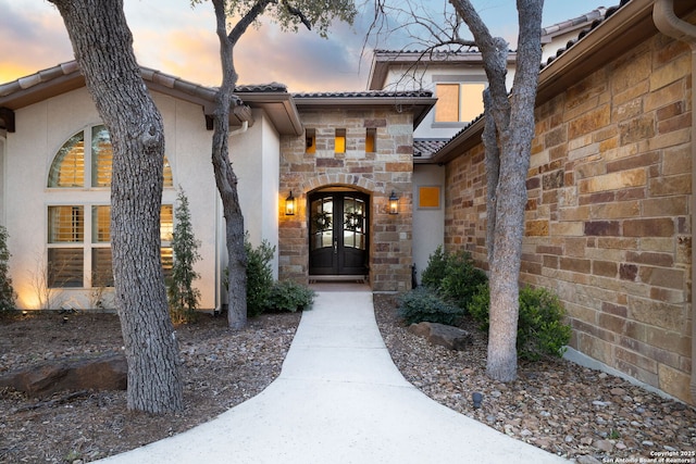 exterior entry at dusk featuring stone siding, french doors, a tiled roof, and stucco siding