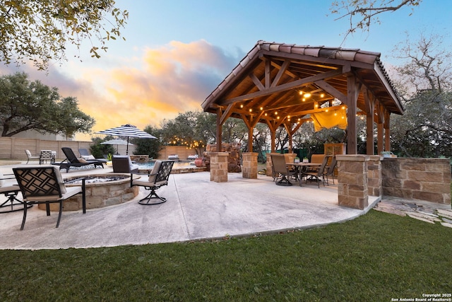 patio terrace at dusk with fence, a fire pit, a gazebo, and a lawn