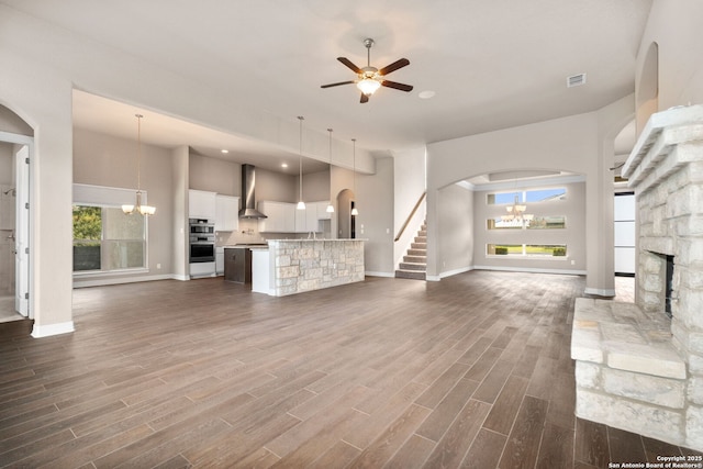 unfurnished living room featuring visible vents, dark wood finished floors, stairway, a fireplace, and ceiling fan with notable chandelier