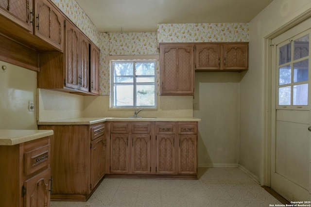 kitchen with wallpapered walls, brown cabinetry, light countertops, light floors, and a sink