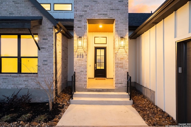 exterior entry at dusk featuring a shingled roof, board and batten siding, and brick siding