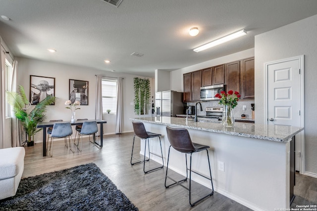 kitchen with light wood finished floors, appliances with stainless steel finishes, a breakfast bar, a kitchen island with sink, and dark brown cabinets