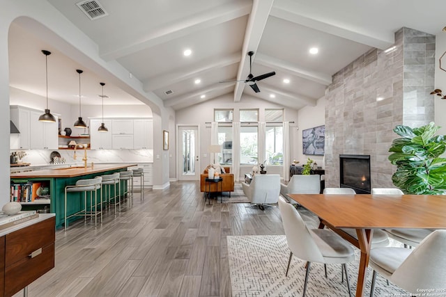 dining space featuring beam ceiling, visible vents, a tiled fireplace, light wood-style floors, and ceiling fan