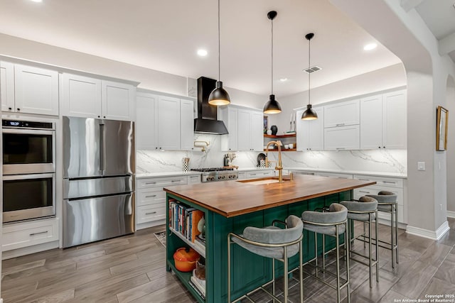 kitchen with open shelves, wooden counters, appliances with stainless steel finishes, a sink, and wall chimney range hood