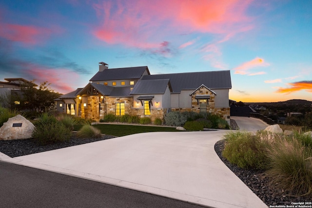 modern farmhouse with metal roof, stone siding, driveway, a standing seam roof, and a chimney