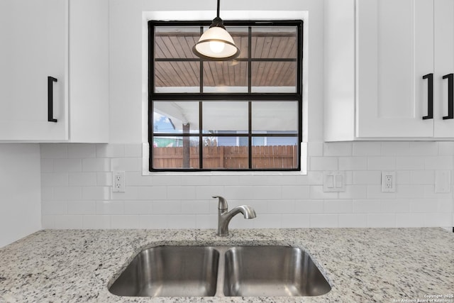 kitchen featuring light stone counters, a sink, white cabinetry, backsplash, and decorative light fixtures