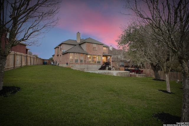 back of property at dusk featuring a yard, a fenced backyard, a chimney, and brick siding