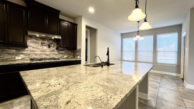 kitchen with light stone counters, under cabinet range hood, stainless steel gas cooktop, a sink, and decorative backsplash