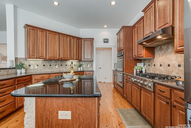 kitchen with stainless steel appliances, dark countertops, brown cabinetry, and under cabinet range hood