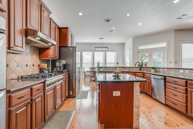 kitchen with appliances with stainless steel finishes, brown cabinetry, a kitchen island, a sink, and under cabinet range hood