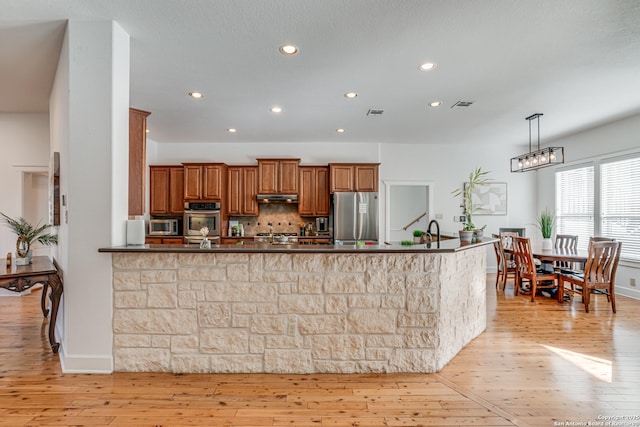 kitchen featuring stainless steel appliances, dark countertops, light wood-style flooring, brown cabinetry, and under cabinet range hood