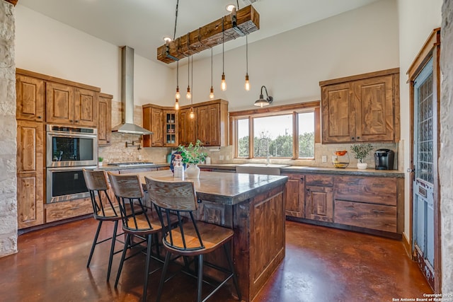 kitchen featuring a towering ceiling, appliances with stainless steel finishes, brown cabinetry, wall chimney range hood, and concrete floors