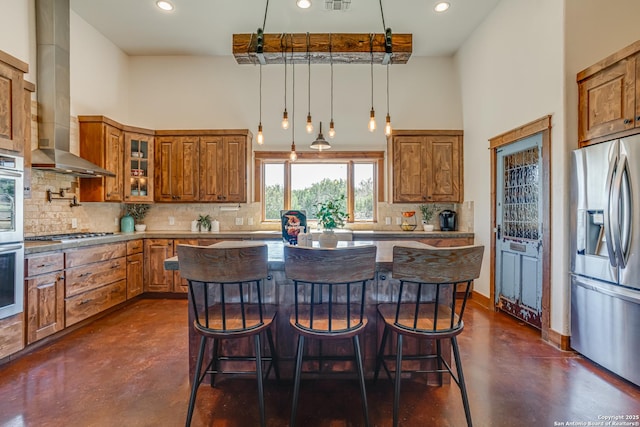 kitchen featuring brown cabinetry, wall chimney exhaust hood, appliances with stainless steel finishes, a high ceiling, and concrete floors