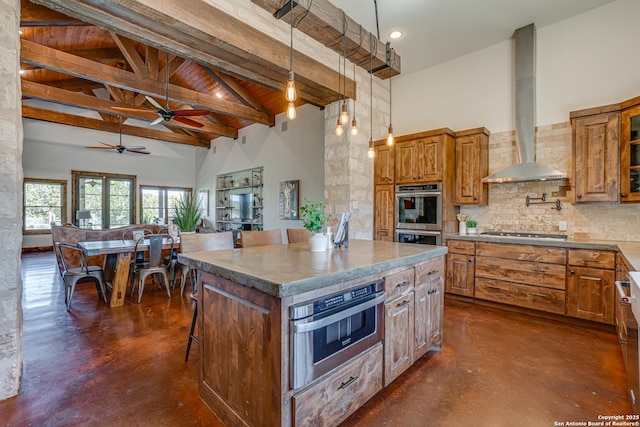 kitchen featuring wall chimney range hood, appliances with stainless steel finishes, brown cabinetry, and high vaulted ceiling