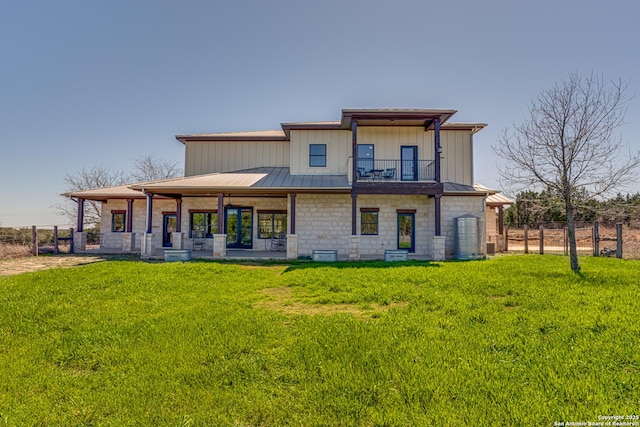 rear view of property with a lawn, a balcony, stone siding, metal roof, and fence