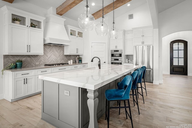 kitchen featuring arched walkways, premium range hood, a sink, appliances with stainless steel finishes, and an island with sink