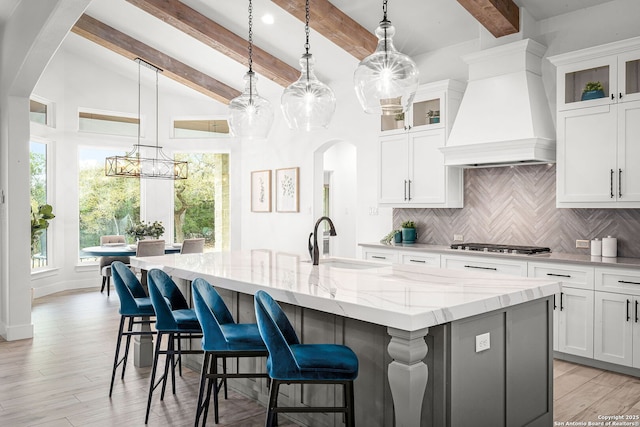 kitchen with arched walkways, premium range hood, a sink, tasteful backsplash, and an island with sink