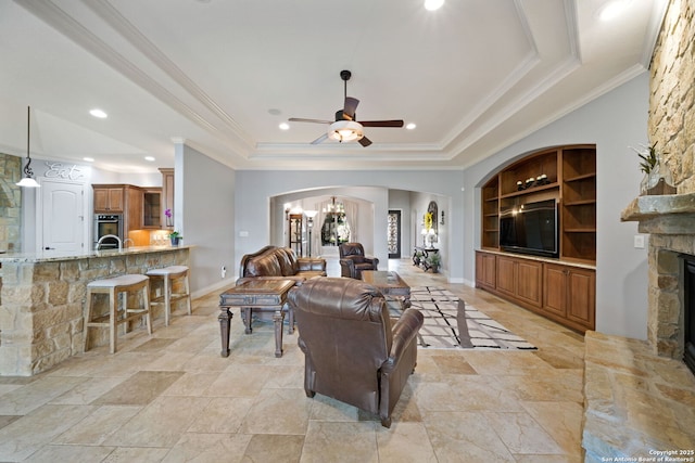 living area featuring arched walkways, a stone fireplace, built in shelves, ornamental molding, and a tray ceiling