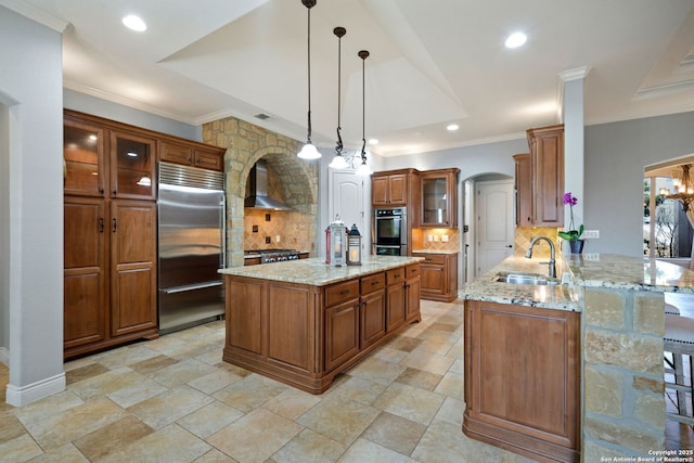 kitchen with arched walkways, a sink, wall chimney range hood, appliances with stainless steel finishes, and brown cabinetry