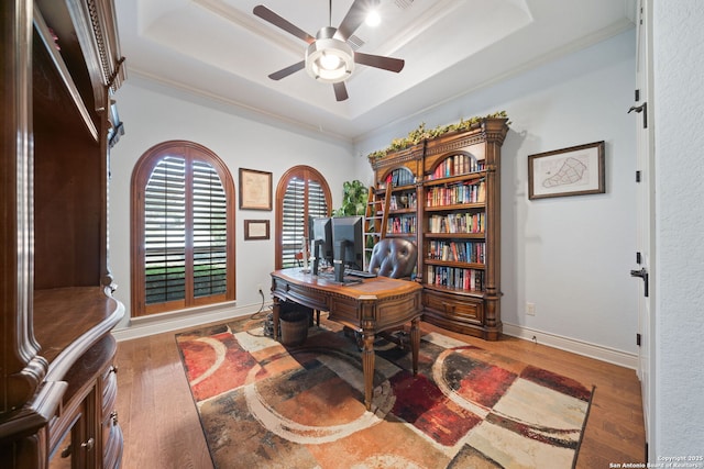 office space with baseboards, a raised ceiling, ceiling fan, wood finished floors, and crown molding