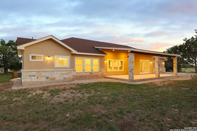 back of house at dusk with stone siding, a patio area, a yard, and stucco siding