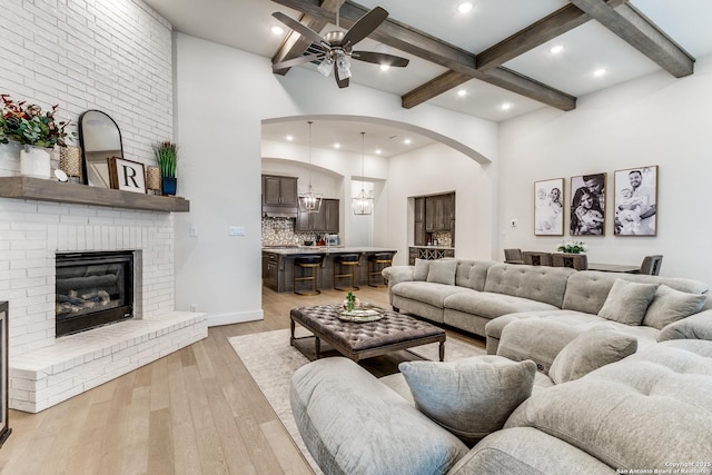 living area featuring light wood-style flooring, a towering ceiling, a ceiling fan, a brick fireplace, and beamed ceiling