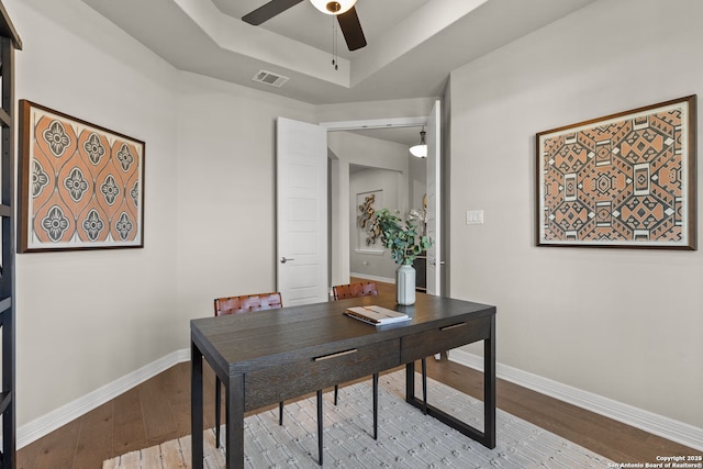 home office with a raised ceiling, visible vents, a ceiling fan, baseboards, and hardwood / wood-style flooring