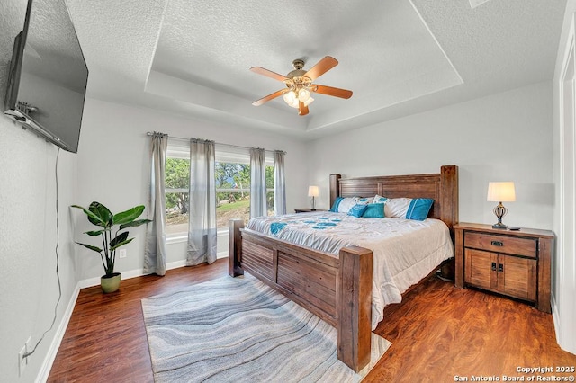 bedroom featuring a raised ceiling, a ceiling fan, a textured ceiling, wood finished floors, and baseboards