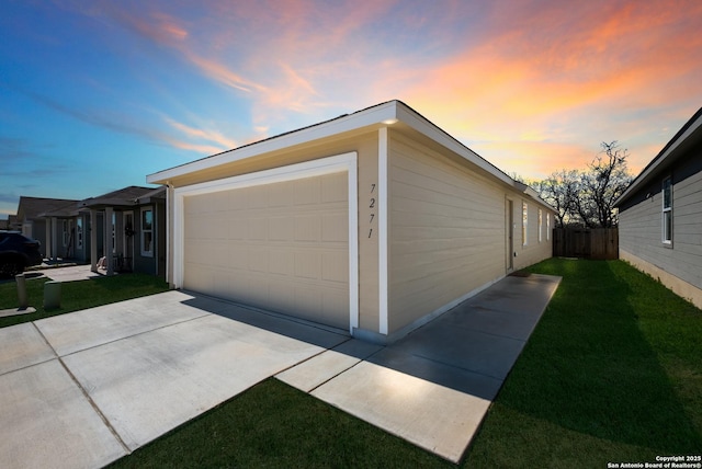 garage at dusk featuring concrete driveway, a lawn, and fence