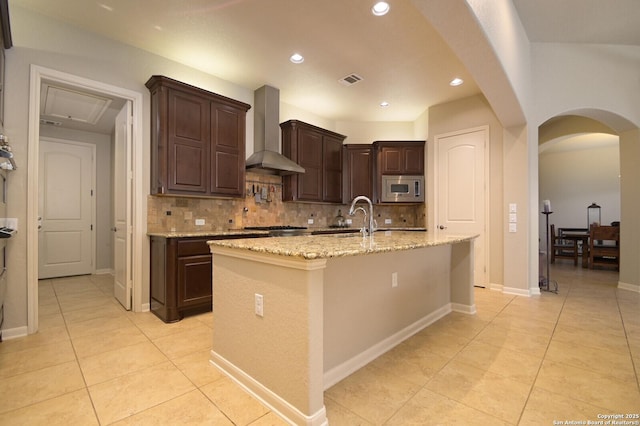 kitchen with arched walkways, a sink, wall chimney exhaust hood, tasteful backsplash, and stainless steel microwave