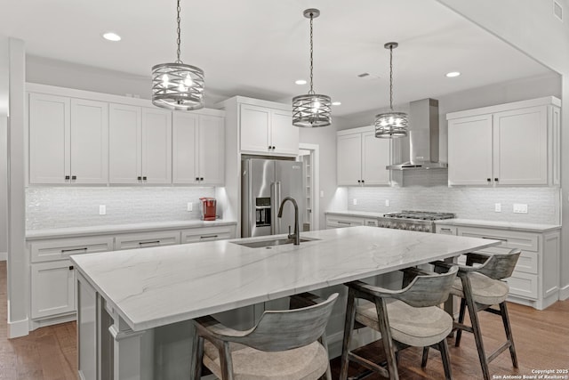 kitchen featuring an island with sink, wall chimney exhaust hood, light wood-style flooring, appliances with stainless steel finishes, and a sink