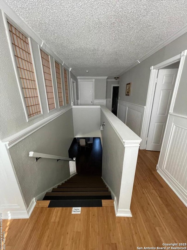 staircase with a textured ceiling, a textured wall, a wainscoted wall, wood finished floors, and crown molding