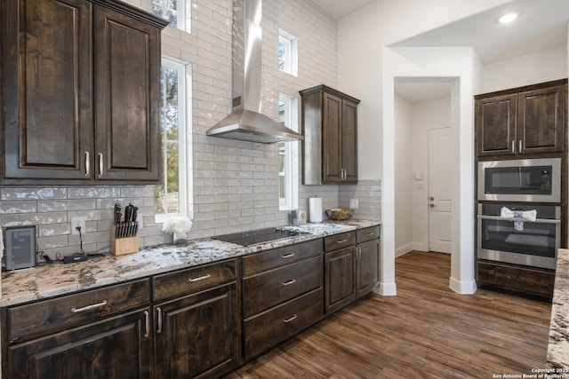 kitchen with dark brown cabinetry, dark wood-style floors, appliances with stainless steel finishes, light stone counters, and wall chimney range hood