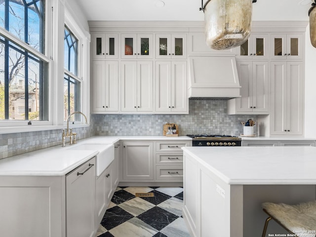 kitchen with tasteful backsplash, glass insert cabinets, premium range hood, light countertops, and a sink