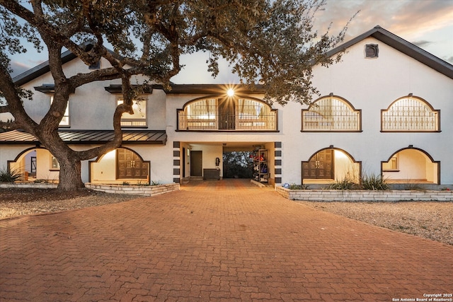 back of property at dusk with a balcony, metal roof, a standing seam roof, decorative driveway, and stucco siding