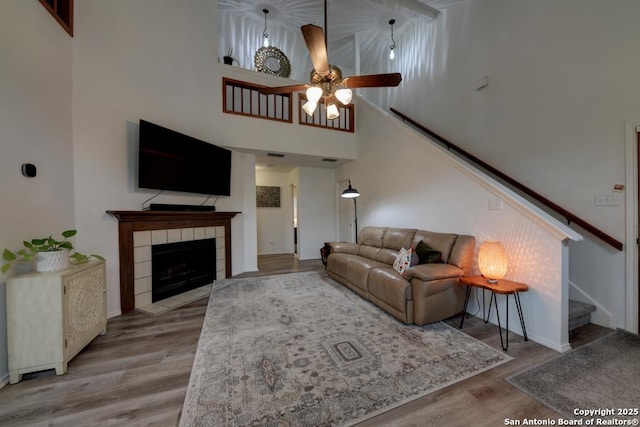 living area with stairs, a ceiling fan, a tiled fireplace, and wood finished floors