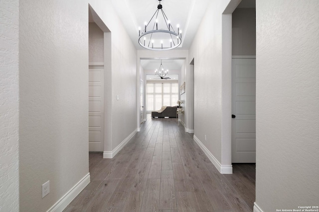 hallway with wood finished floors, a textured wall, baseboards, and an inviting chandelier