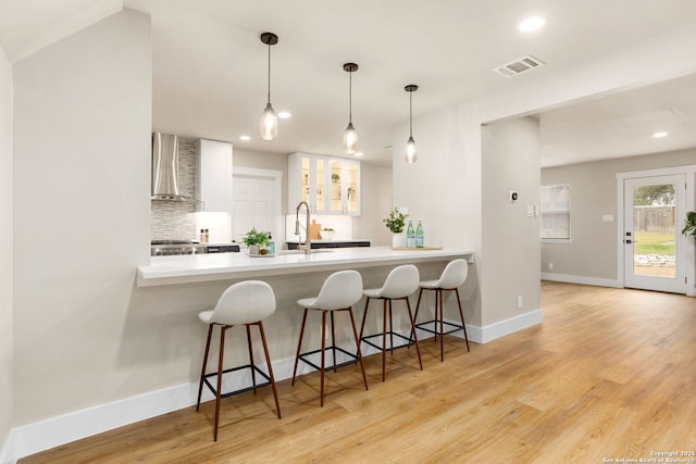 kitchen with a peninsula, a sink, visible vents, wall chimney exhaust hood, and light wood finished floors