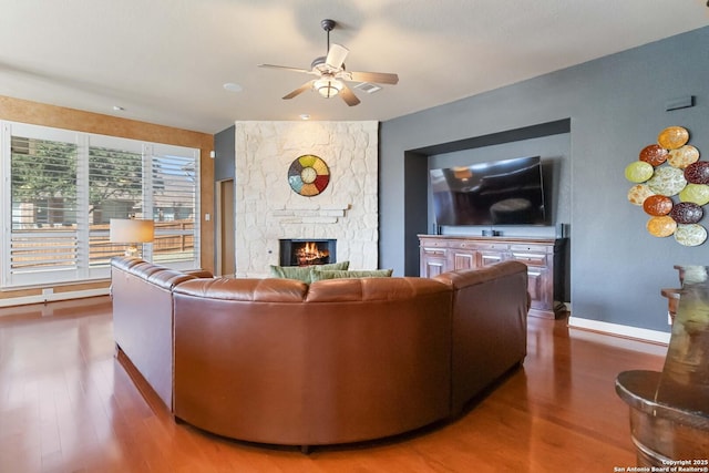 living area featuring ceiling fan, a stone fireplace, baseboards, and wood finished floors