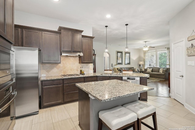 kitchen featuring dark brown cabinetry, tasteful backsplash, light stone countertops, stainless steel appliances, and under cabinet range hood