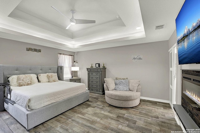 bedroom with visible vents, a tray ceiling, wood finished floors, and a glass covered fireplace