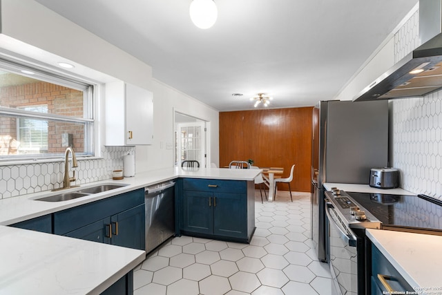 kitchen featuring blue cabinets, a peninsula, stainless steel appliances, wall chimney range hood, and a sink