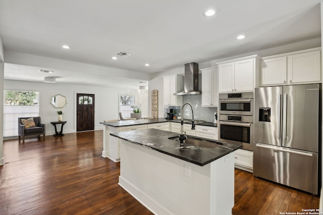 kitchen with wall chimney range hood, a peninsula, visible vents, appliances with stainless steel finishes, and a sink