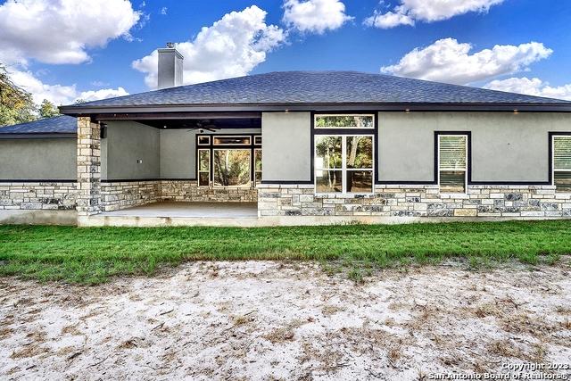 rear view of property featuring a shingled roof, a patio, stone siding, a chimney, and stucco siding