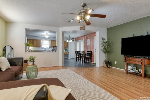 living area featuring ceiling fan with notable chandelier, light wood-style floors, baseboards, and a textured ceiling