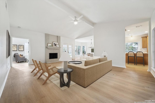 living area featuring beam ceiling, light wood-style flooring, a brick fireplace, baseboards, and ceiling fan with notable chandelier